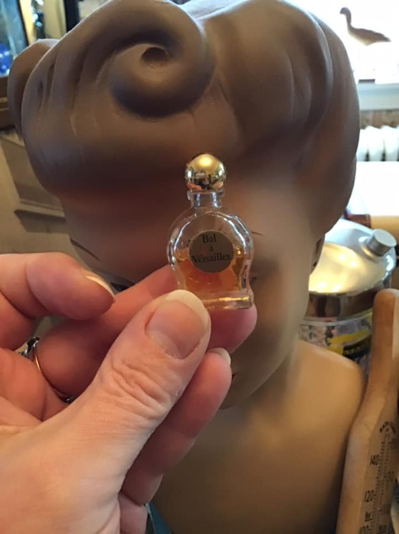 Puede incluir: Un frasco de perfume en miniatura de vidrio transparente con un tap&oacute;n esf&eacute;rico de tono dorado, que contiene un l&iacute;quido de color &aacute;mbar. La etiqueta frontal presenta el texto Bal a Versailles en letras negras sobre el cuerpo redondeado.