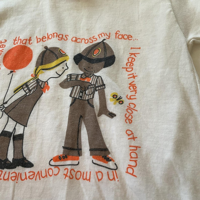 May include: A white t-shirt with a cartoon illustration of two children. The text on the shirt reads "@ that belongs across my face... I keep it very close at hand in a most convenient"