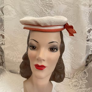 May include: A cream-colored textured beret-style hat featuring a horizontal orange band around the base and a small orange ribbon bow detail on the right side. The hat is displayed on a mannequin head with brown hair and painted red lips.