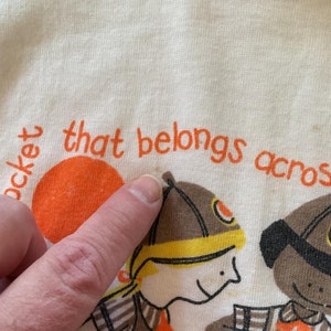 May include: A white t-shirt with orange text that reads "pocket that belongs across". The shirt features two cartoon characters, one wearing a brown hat and the other wearing a black hat.