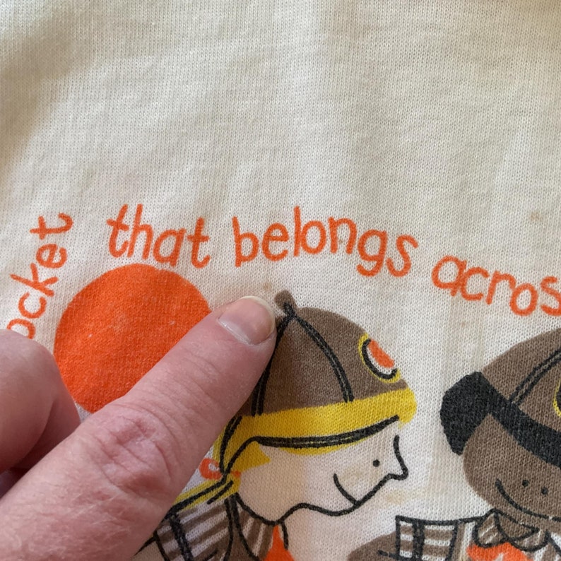 May include: A white t-shirt with orange text that reads "pocket that belongs across". The shirt features two cartoon characters, one wearing a brown hat and the other wearing a black hat.