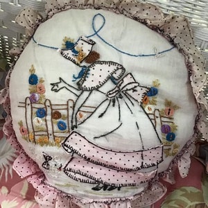 K&ouml;nnte beinhalten: Ein rundes, wei&szlig;es Kissen mit einem rosa und schwarz gepunkteten Volant. Das Kissen ist mit einer Frau in einem rosa Kleid und einem wei&szlig;en Hut bestickt, die eine Schnur mit einem blauen Vogel am Ende h&auml;lt. Die Frau steht vor einem Zaun mit Blumen.