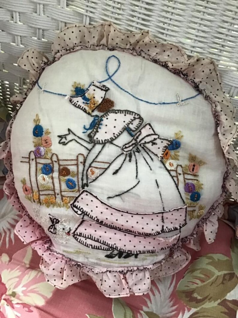 K&ouml;nnte beinhalten: Ein rundes, wei&szlig;es Kissen mit einem rosa und schwarz gepunkteten Volant. Das Kissen ist mit einer Frau in einem rosa Kleid und einem wei&szlig;en Hut bestickt, die eine Schnur mit einem blauen Vogel am Ende h&auml;lt. Die Frau steht vor einem Zaun mit Blumen.