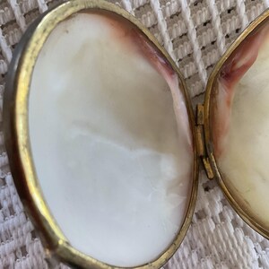 May include: Open oval-shaped locket with a gold-colored frame and a white interior with pink and red accents. The locket is hinged and appears to be empty. The locket is on a white, textured surface.
