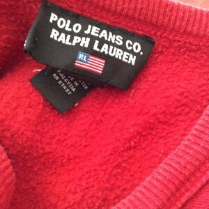 May include: A black tag with white text that reads "Polo Jeans Co. Ralph Lauren" with a small American flag and the letters "RL" in the upper right corner. The tag is attached to a red sweatshirt.