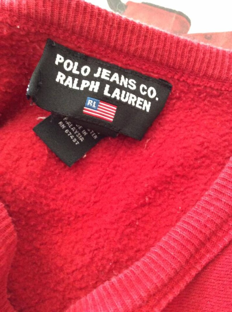 May include: A black tag with white text that reads "Polo Jeans Co. Ralph Lauren" with a small American flag and the letters "RL" in the upper right corner. The tag is attached to a red sweatshirt.