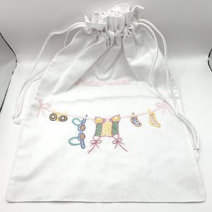 May include: White drawstring bag with embroidered underwear design. The bag features a string of colorful embroidered underwear, including socks, a bra, and a pair of underwear. The word "underwear" is embroidered in pink on the bag.