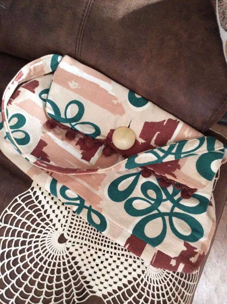 May include: A small, cream-colored fabric bag with a green and brown abstract pattern. The bag has a brown tassel and a wooden button closure.