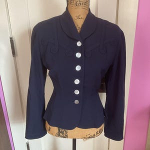 May include: A navy blue blazer with a fitted waist and long sleeves. The blazer has a pointed collar and a row of white buttons down the front. The shoulders are slightly padded. The blazer has decorative stitching around the collar and pockets.