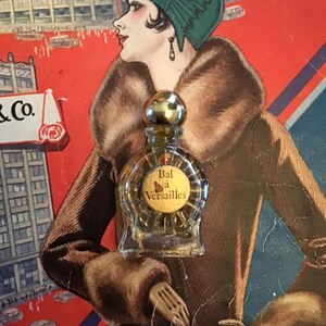 Puede incluir: Un peque&ntilde;o frasco de perfume de vidrio transparente con un tap&oacute;n esf&eacute;rico dorado y una etiqueta redonda con el texto Bal &agrave; Versailles. El frasco est&aacute; colocado sobre una ilustraci&oacute;n de estilo vintage de una persona con sombrero verde.