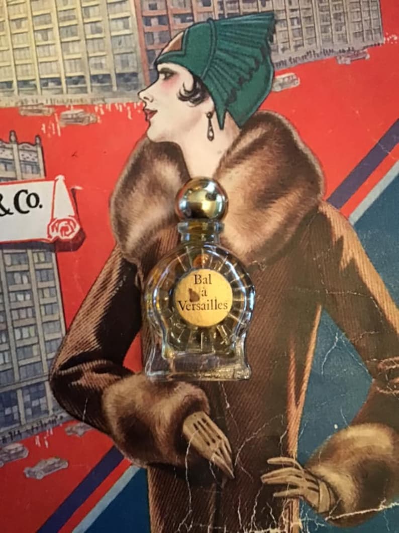 Puede incluir: Un peque&ntilde;o frasco de perfume de vidrio transparente con un tap&oacute;n esf&eacute;rico dorado y una etiqueta redonda con el texto Bal &agrave; Versailles. El frasco est&aacute; colocado sobre una ilustraci&oacute;n de estilo vintage de una persona con sombrero verde.