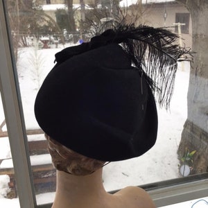 Puede incluir: Un sombrero de fieltro negro con una gran pluma negra en el lateral. El sombrero est&aacute; en una cabeza de maniqu&iacute;.