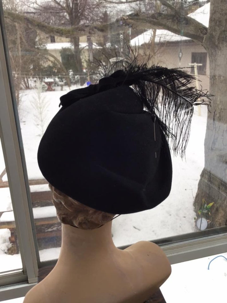 Puede incluir: Un sombrero de fieltro negro con una gran pluma negra en el lateral. El sombrero est&aacute; en una cabeza de maniqu&iacute;.
