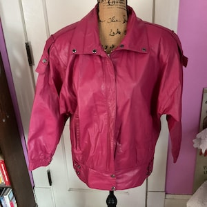May include: A vibrant fuchsia jacket with a classic design. The jacket features a stand-up collar, shoulder epaulets, and a zip-up front. The sleeves are three-quarter length, and the jacket has a slightly cropped hemline. Silver-toned snaps and details add a touch of contrast.