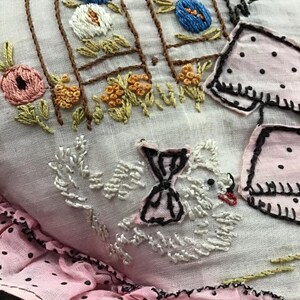 K&ouml;nnte beinhalten: Ein wei&szlig;es Stoffkissen mit rosa Punkten am Rand. Das Kissen ist mit einem wei&szlig;en Vogel mit schwarzer Schleife, einem braunen Zaun mit Blumen und einem rosa-wei&szlig;en Kleid mit schwarzen Punkten bestickt.
