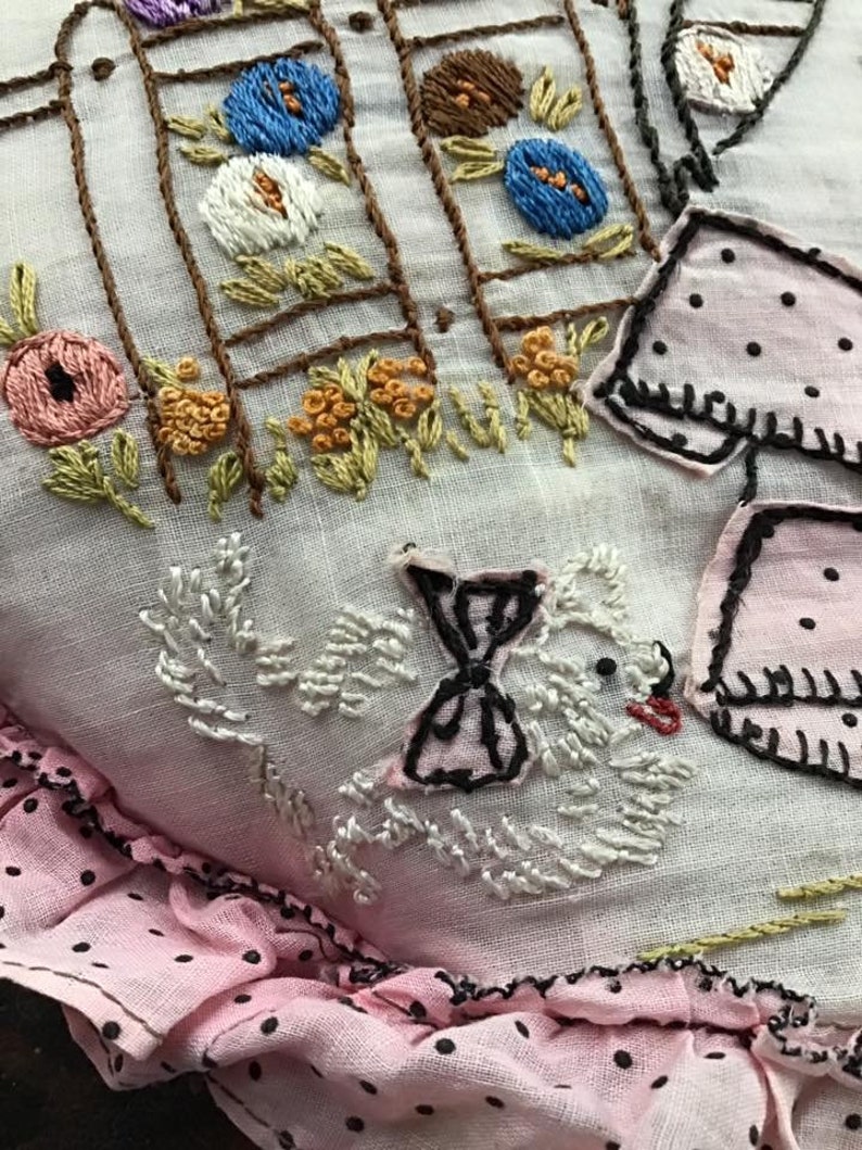 K&ouml;nnte beinhalten: Ein wei&szlig;es Stoffkissen mit rosa Punkten am Rand. Das Kissen ist mit einem wei&szlig;en Vogel mit schwarzer Schleife, einem braunen Zaun mit Blumen und einem rosa-wei&szlig;en Kleid mit schwarzen Punkten bestickt.