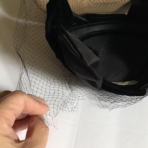 Puede incluir: Sombrero de terciopelo negro con velo de red negro. El sombrero tiene una forma redondeada y una textura suave y aterciopelada.