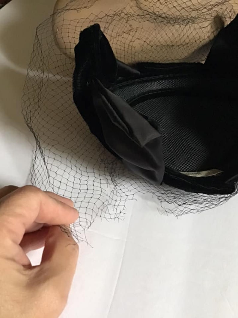Puede incluir: Sombrero de terciopelo negro con velo de red negro. El sombrero tiene una forma redondeada y una textura suave y aterciopelada.