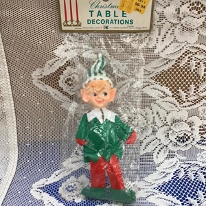 Op de afbeelding: Een vintage kersttafeldecoratie met een groene en witte elf met rode broek. De elf is verpakt in plastic en heeft een prijskaartje met de tekst "Christmas Table Decorations, Key 1 98-94 38c".