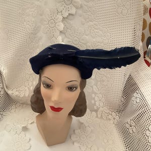 Puede incluir: Un gorro azul marino de fieltro con un adorno de pluma. El gorro está colocado en una cabeza de maniquí con cabello castaño.