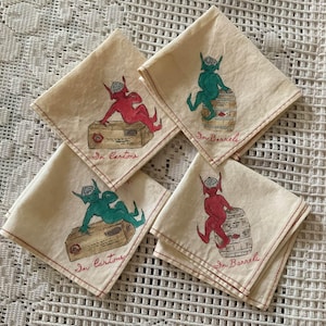 May include: Four off-white fabric napkins with red stitched edges. Each features a hand-drawn illustration of a small creature wearing a hat. Two napkins show a red or green creature on a box labeled In Cartons, and two show a creature on a barrel labeled In Barrels.