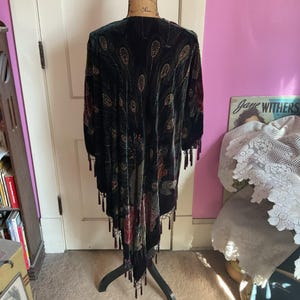 Puede incluir: Un chal de terciopelo verde oscuro con un diseño de plumas de pavo real, adornado con cuentas y borlas. El chal se exhibe en un maniquí, mostrando su forma fluida y sus intrincados detalles. El diseño incluye acentos florales.