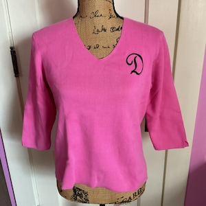 Könnte beinhalten: Ein leuchtend pinkfarbener V-Ausschnitt-Pullover mit Dreiviertelärmeln. Der Pullover hat einen schwarz gestickten Buchstaben "D" auf der linken Brust. Das Kleidungsstück wird auf einer Schaufensterpuppe präsentiert.