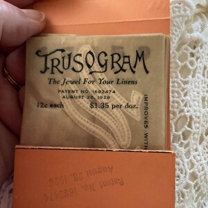 May include: A vintage tan paper packet labeled Trusogram The Jewel For Your Linens with patent number 1682474 dated August 28, 1928. The packet displays pricing of 12c each and $1.35 per doz, featuring a decorative embroidered letter design.