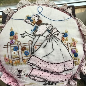 K&ouml;nnte beinhalten: Ein vintage besticktes Kissen mit einem rosa-wei&szlig;en Polka-Dot-Stoff. Die Stickerei zeigt eine Frau in einem wei&szlig;en Kleid mit einem rosa-wei&szlig;en Polka-Dot-Rock, die eine Schnur mit einem blauen Vogel am Ende h&auml;lt. Die Frau steht in einem Garten mit Blumen und einem Zaun.