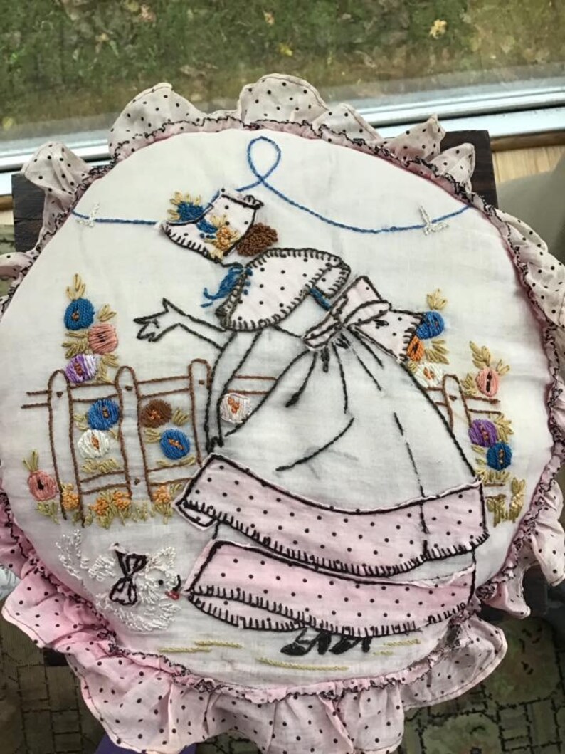 K&ouml;nnte beinhalten: Ein vintage besticktes Kissen mit einem rosa-wei&szlig;en Polka-Dot-Stoff. Die Stickerei zeigt eine Frau in einem wei&szlig;en Kleid mit einem rosa-wei&szlig;en Polka-Dot-Rock, die eine Schnur mit einem blauen Vogel am Ende h&auml;lt. Die Frau steht in einem Garten mit Blumen und einem Zaun.