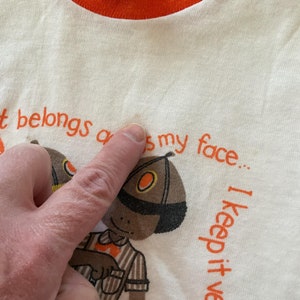 May include: White t-shirt with an orange ringer neck. The shirt has a cartoon illustration of a person wearing a brown and orange hat and a striped shirt. The text on the shirt reads "It belongs on my face... I keep it well-dressed."