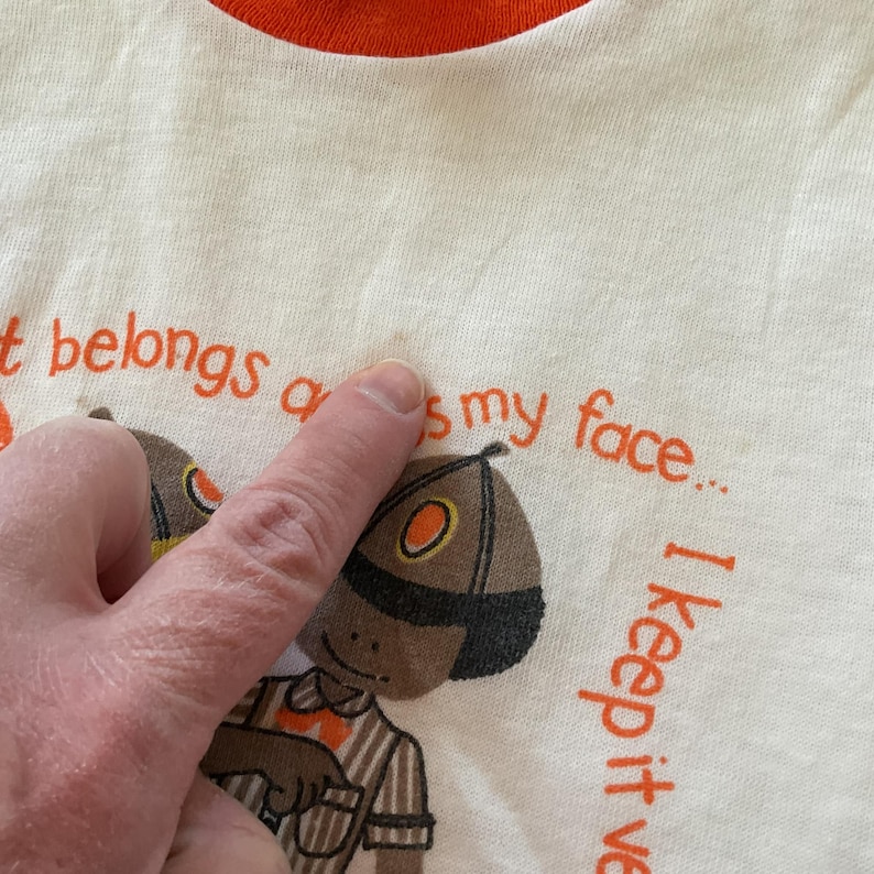 May include: White t-shirt with an orange ringer neck. The shirt has a cartoon illustration of a person wearing a brown and orange hat and a striped shirt. The text on the shirt reads "It belongs on my face... I keep it well-dressed."