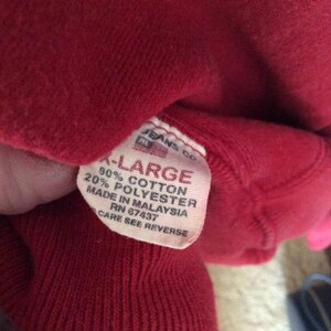 May include: A red sweatshirt tag with white text that reads "Jeans Co. -Large 90% Cotton 20% Polyester Made in Malaysia RN 87437 Care See Reverse"