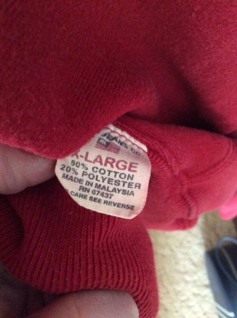 May include: A red sweatshirt tag with white text that reads "Jeans Co. -Large 90% Cotton 20% Polyester Made in Malaysia RN 87437 Care See Reverse"