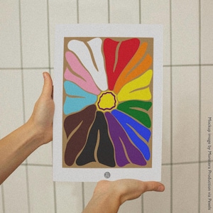 May include: A colorful floral art print featuring a large flower with petals in various colors, including red, orange, yellow, and purple. The flower is centered on a tan background, framed by a white border. The art print is held by hands.