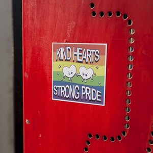 Könnte beinhalten: Ein Aufkleber mit dem Text "KIND HEARTS STRONG PRIDE" in Weiß. Er zeigt zwei Cartoon-Herzen, die sich an den Händen halten, vor einem Regenbogenhintergrund. Der Aufkleber befindet sich auf einer roten Oberfläche.
