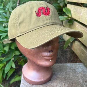 Könnte beinhalten: Olivgrüne Baseballkappe mit gebogenem Schirm. Die Kappe hat ein rotes, gesticktes Design einer Raupe auf der Vorderseite. Die Kappe wird auf einem Schaufensterkopf präsentiert.