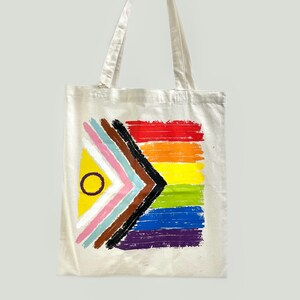 Könnte beinhalten: Cremefarbene Tragetasche mit einem farbenfrohen, gemalten Design. Das Design umfasst einen Regenbogen aus horizontalen Streifen und eine geometrische Form mit einem gelben Dreieck und einem schwarzen Kreis. Die Tasche hat zwei Griffe.