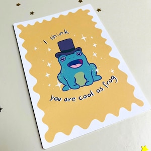 Könnte beinhalten: Eine Grußkarte mit einem Cartoon-Frosch mit Zylinder. Der Frosch ist blau mit grünem Bauch und offenem Mund. Die Karte hat den Text "I think You are cool as frog" auf einem gelben Hintergrund mit gewelltem Rand.