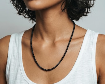 Puede incluir: Un collar compuesto por pequeñas cuentas esféricas de color negro mate y uniforme, ensartadas en un diseño circular continuo. Las cuentas son lisas y oscuras, creando un accesorio monocromático sencillo que descansa sobre el cuello.