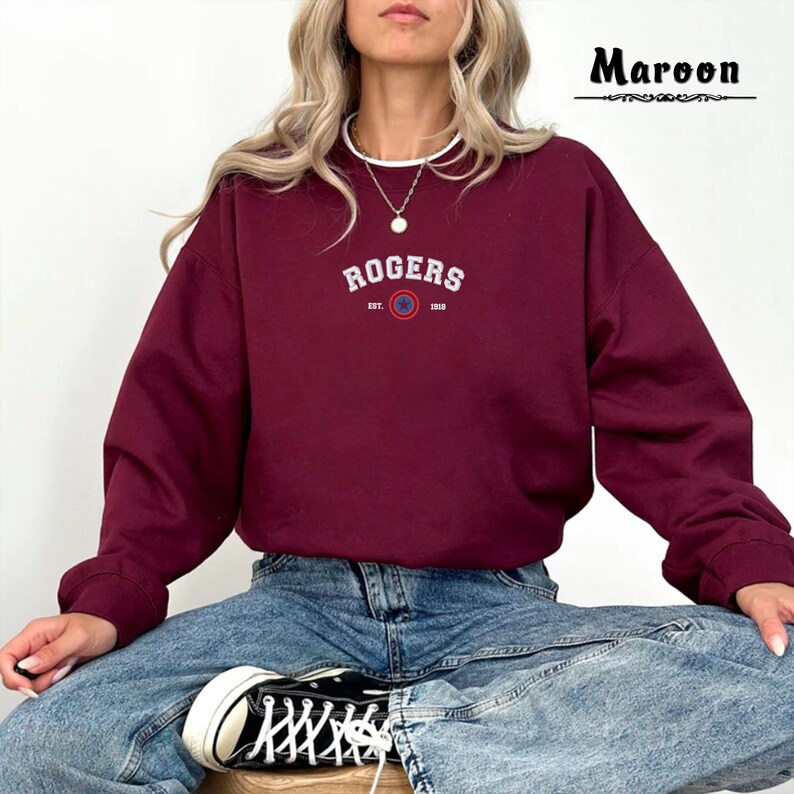 Może przedstawiać: Bordowa bluza z napisem "ROGERS EST. 1910" i logo w kolorze białym i niebieskim.