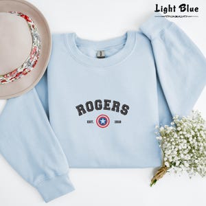 Może przedstawiać: Jasno błękitna bluza z haftowanym czarnym wzorem tarczy ze gwiazdą i tekstem "ROGERS EST. 1918".
