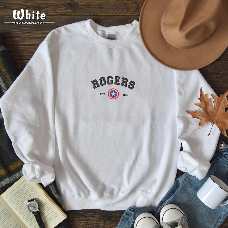Może przedstawiać: Biała bluza z czarnym nadrukiem tekstu "ROGERS" z logo tarczy Kapitana Ameryki w kolorze czerwonym, białym i niebieskim. Tekst "EST. 1918" znajduje się pod logo.