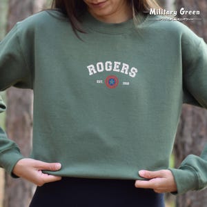 Może przedstawiać: Zielona bluza z kapturem w kolorze khaki z napisem "ROGERS EST. 1918" i czerwoną, białą i niebieską gwiazdą w k&oacute;łku.