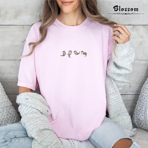 May include: A light pink t-shirt with a small embroidered design of four otters. The otters are in various poses, and the shirt has short sleeves. The word "Blossom" is printed in black script in the upper right corner.