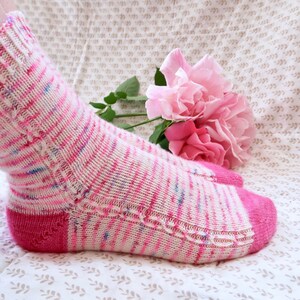 May include: A pair of pink and white striped socks with a pink heel and toe. The socks are knitted with a delicate lace pattern. The socks are lying on a white fabric with a floral pattern.