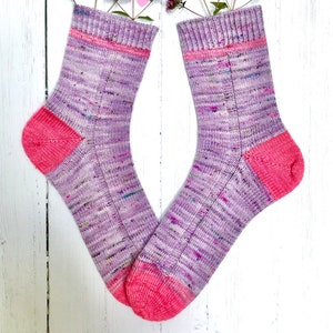 May include: A pair of purple and pink knitted socks with a speckled pattern. The socks are arranged in a V-shape with pink hearts scattered around them.