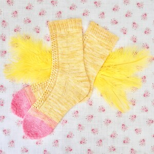 May include: A pair of yellow and pink knitted socks with a delicate lace pattern. The socks are laid out on a floral fabric background.