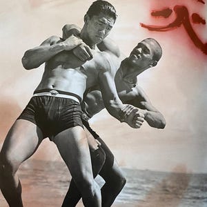 May include: Black and white photograph of two men wrestling on a beach. One man is wearing black shorts with a white waistband. The other man is shirtless. The background shows the ocean and a red Japanese character.