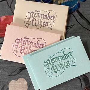 May include: Three rectangular folded cards in cream, light pink, and light blue. Each card features the text "Remember When" printed in a decorative, calligraphic font. The cream card has dark purple text, the pink card has dark pink text, and the light blue card has dark green text.